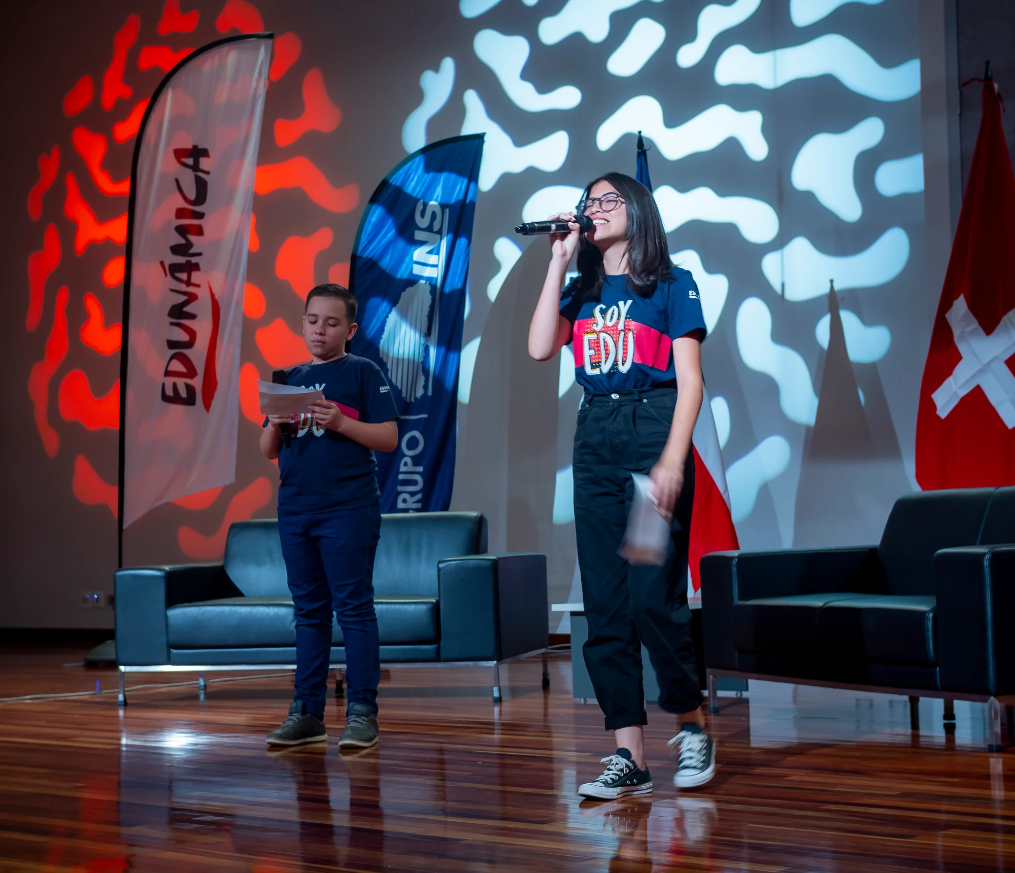 Los dos jóvenes anfitriones de Edunámica presentando en tarima, con las banderas de Costa Rica y Suiza de fondo