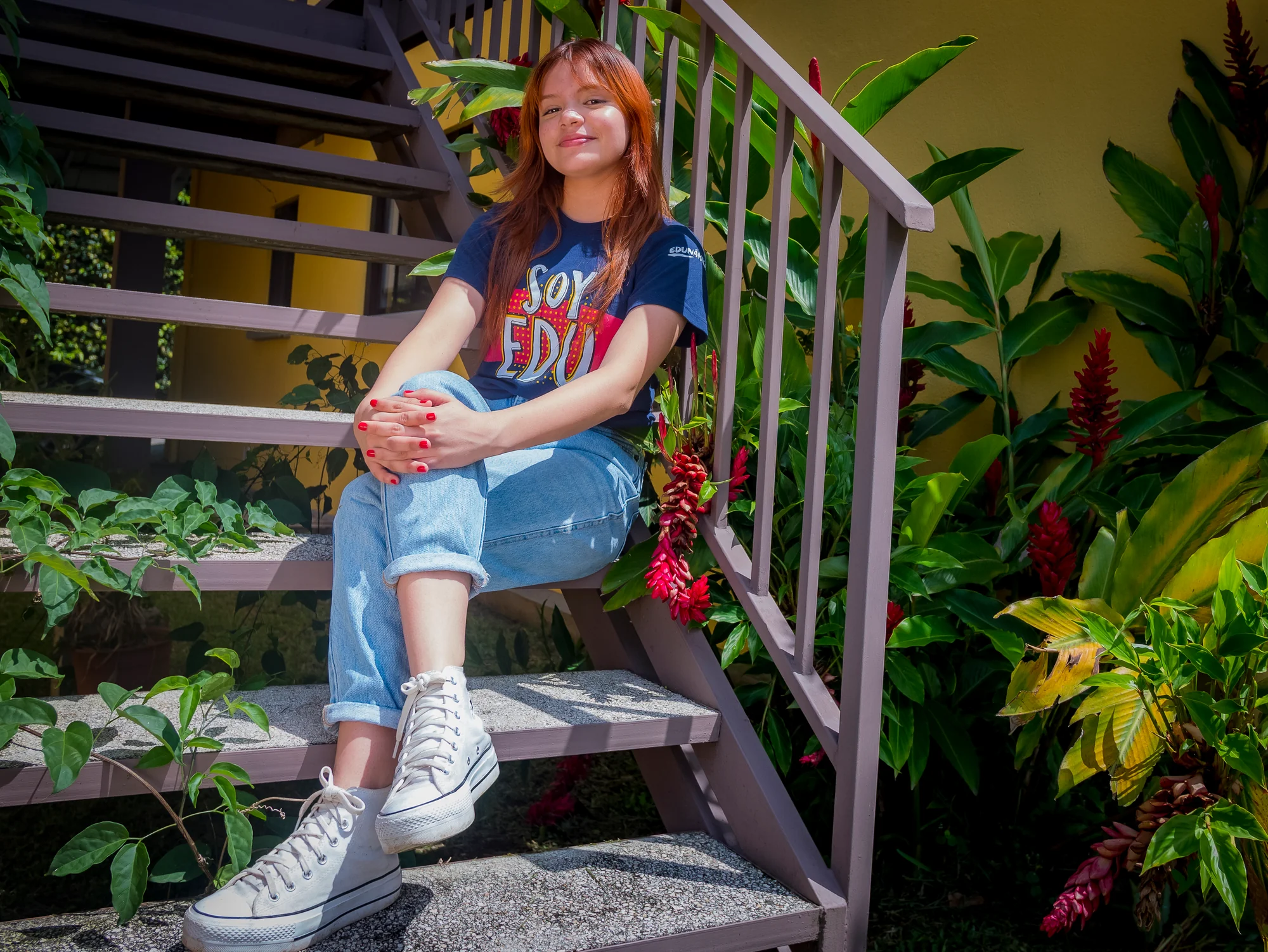 Beneficiaria de Edunámica sentada en una escalera exterior con la camiseta de la campaña Soy Edunámica, rodeada de plantas tropicales